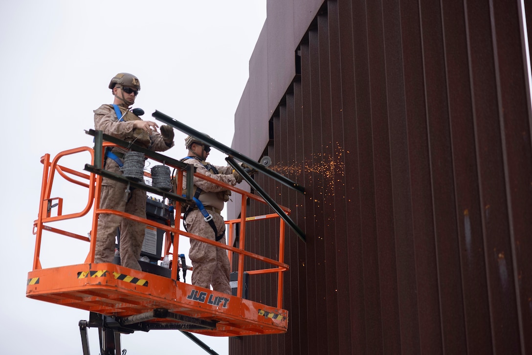 Two Marines standing on an elevated platform use tools to install stakes on a tall metal wall.