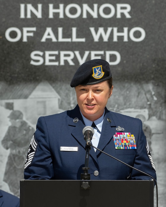 A woman in uniform, wearing a black beret, speaks at a podium in front of a sign that says "In honor of all who serve."