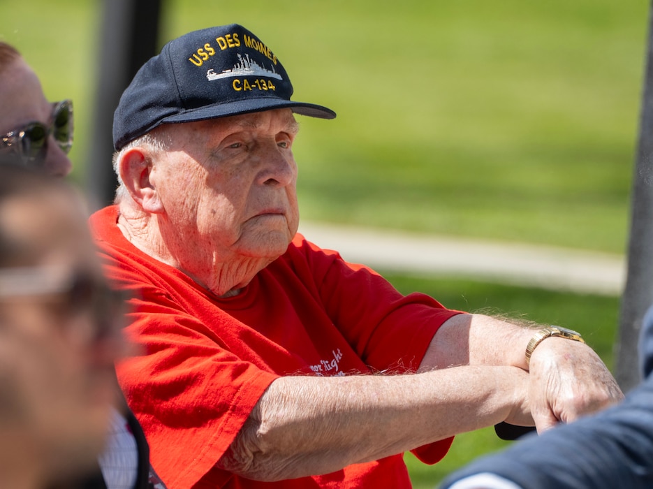 An elderly man in a red tee-shirt and a blue ball cap that says "USS Des Moines, CA-134" sits in the sunshine listening.