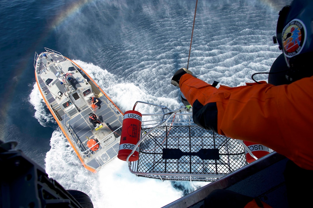A partially visible Coast Guardsman lowers a basket from an airborne helicopter to a boat in wavy waters as a rainbow is seen to the left.