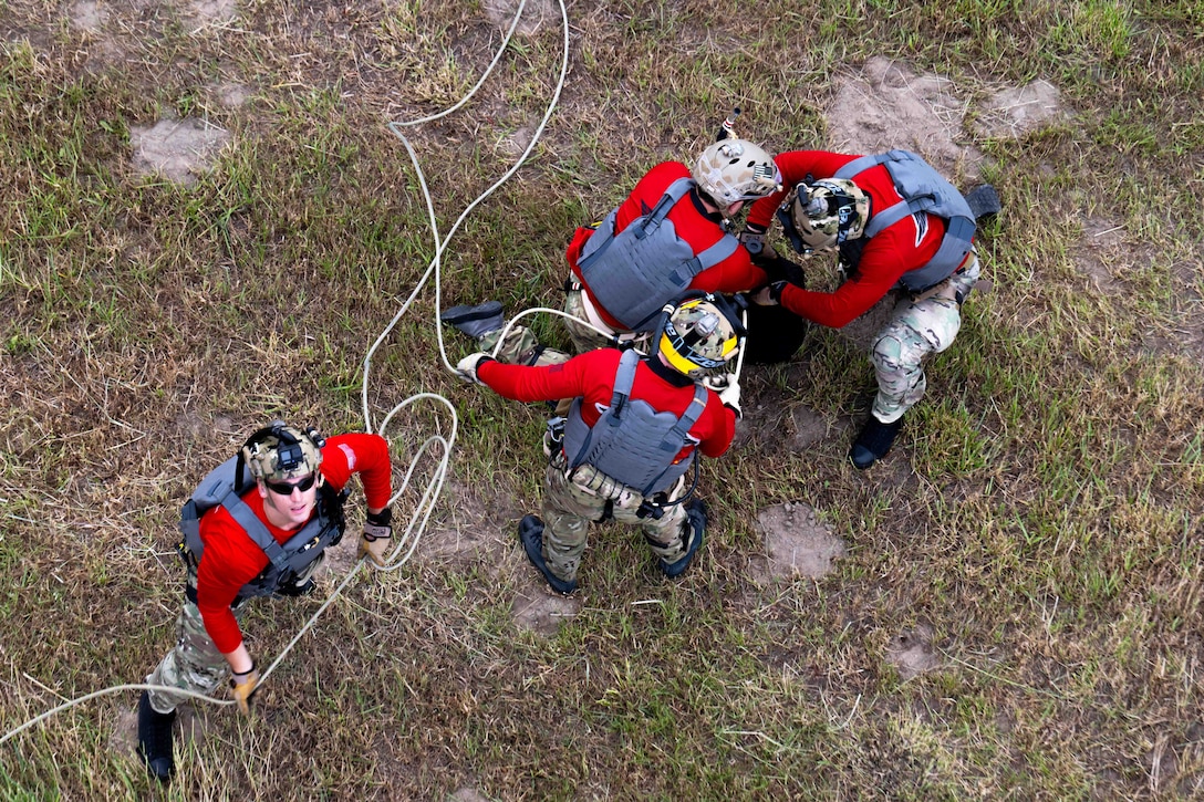 Four airmen in red shirts and tactical gear hold a rope while standing or kneeling in a field as seen from above.