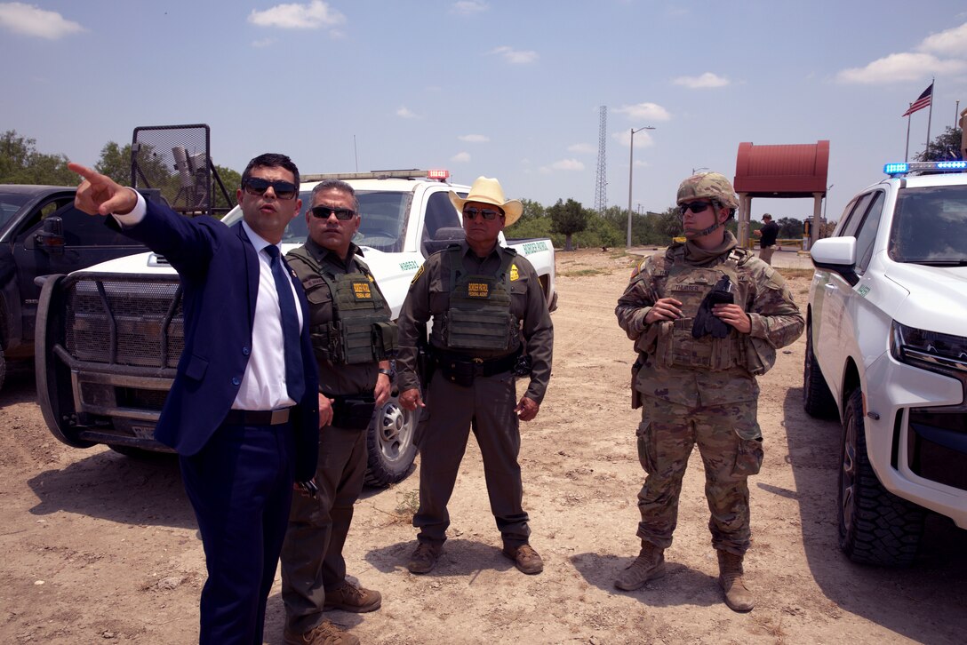 Four people, one wearing a dress blazer and tie and pointing as three others in uniform look into the distance, stand between vehicles on a dirt road.