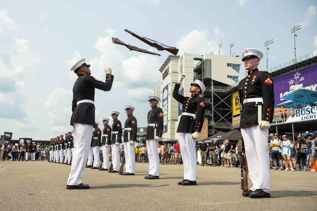 Marines in ceremonial dress stand in formation as two fellow Marines throw rifles in the air as a crowd watches outside of a stadium.