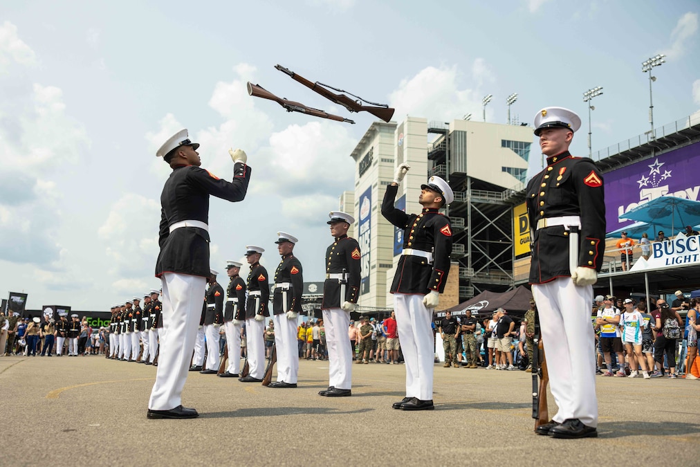 Marines in ceremonial dress stand in formation as two fellow Marines throw rifles in the air as a crowd watches outside of a stadium.