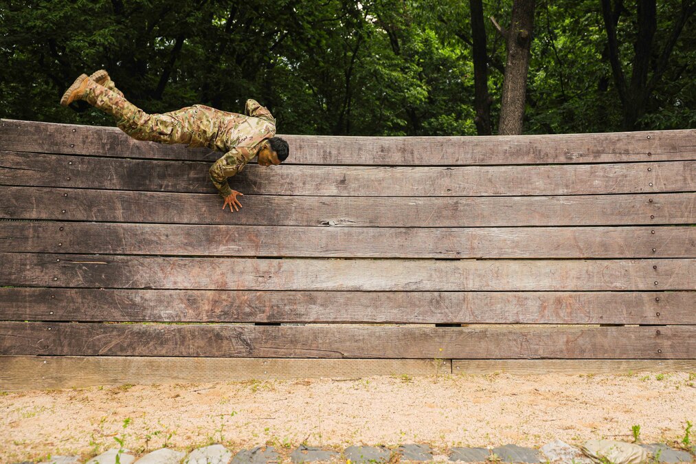 A soldier jumps over a wooden wall obstacle outside.
