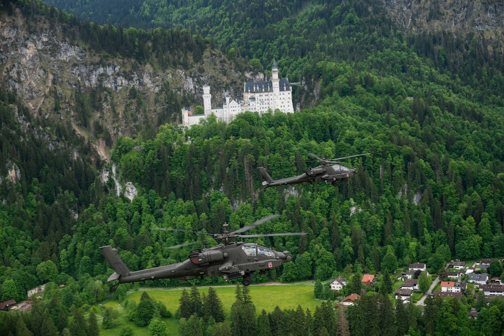 Two Army helicopters fly above a green forest with a castle in the background.