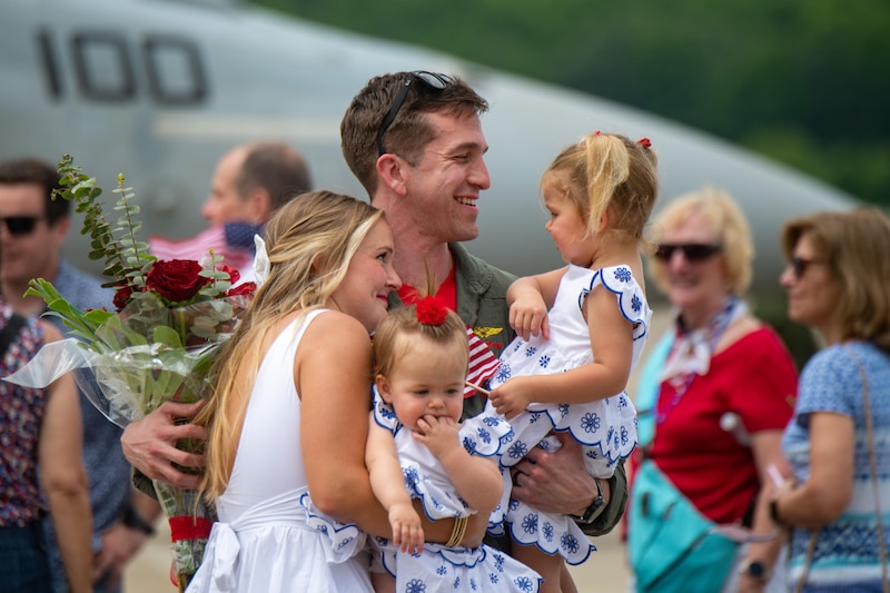 A sailor holds two children and a bouquet while a person hugs all of them.