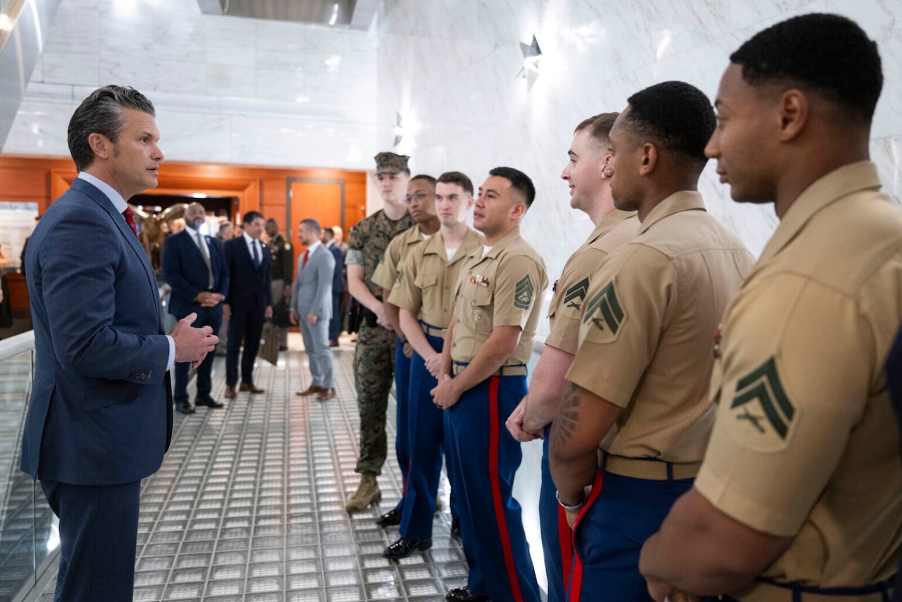 A man in a suit on the left talks to a group of men in military uniforms on the right. Three men in business attire are speaking in the background.