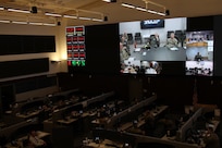 photo of a large room with cubicles and people working and a large projector with multiple screens.