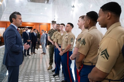 A man in a suit on the left talks to a group of men in military uniforms on the right. Three men in business attire are speaking in the background.