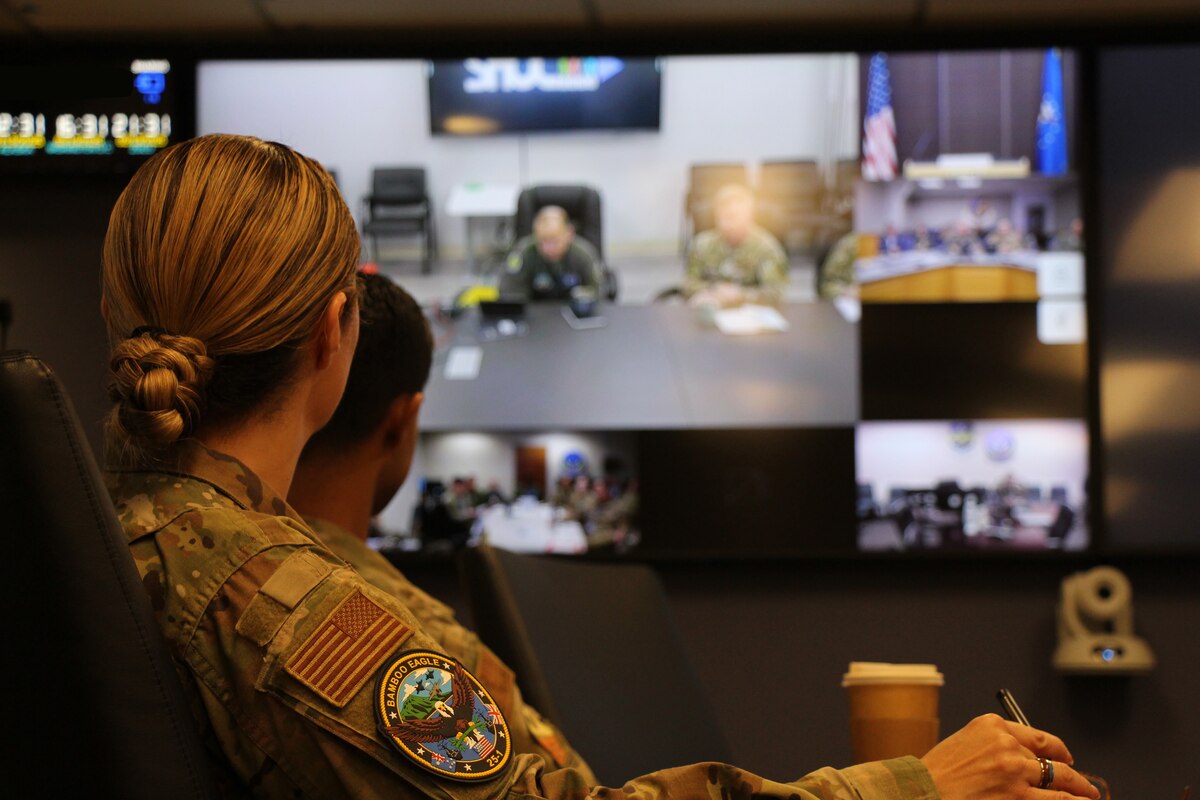 two uniformed U.S. Air Force Airmen listen to a briefing on a large projector