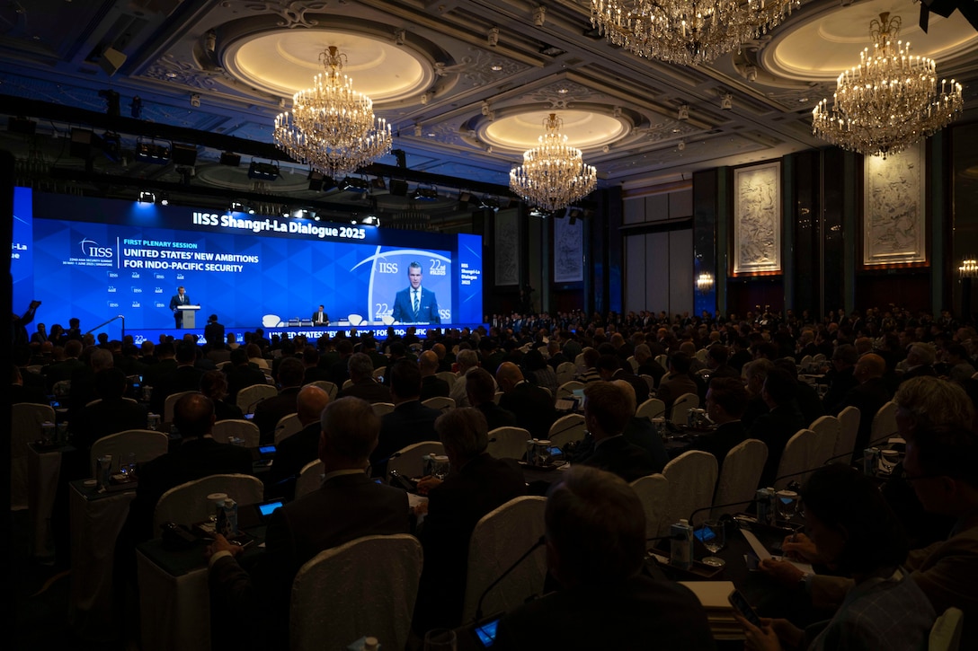 A full audience is seen in a large, darkened room with a speaker at a lectern and under lights on a stage and reflected in a monitor adjacent, all framed in a blue background.