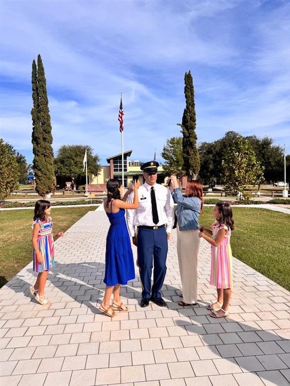Wife and daughters of male Soldier pin his rank during his promotion ceremony. Ceremony is outside on a sunny day.