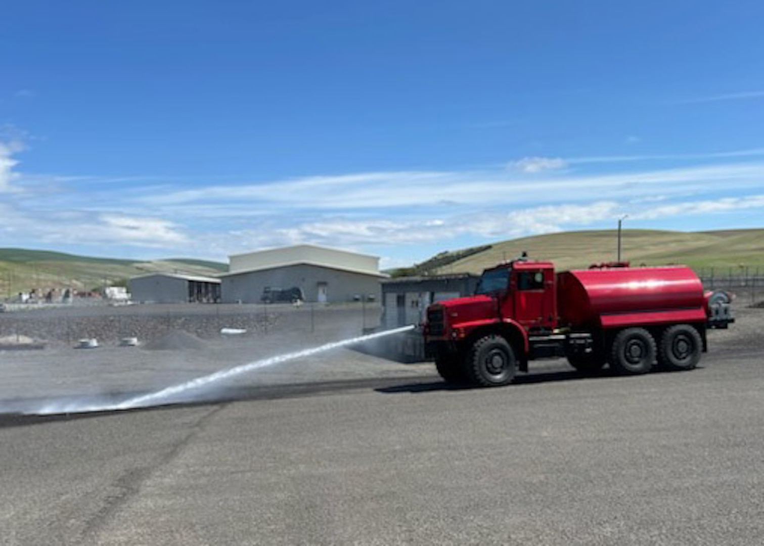 A former military truck now painted red with a large tank on the bed of the truck sprays water from the nose of the truck.