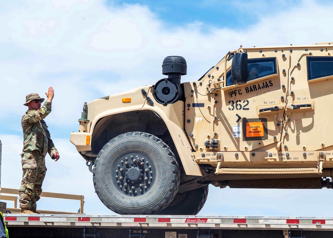 A soldier standing on a trailer uses hand motions to guide a person driving an all-terrain vehicle onto the trailer.