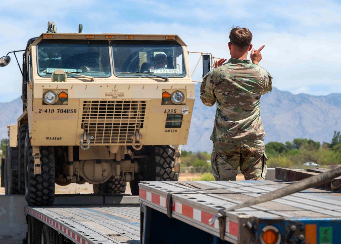 A soldier standing on a vehicle trailer uses hand motions to guide a person driving a tactical truck onto the trailer.
