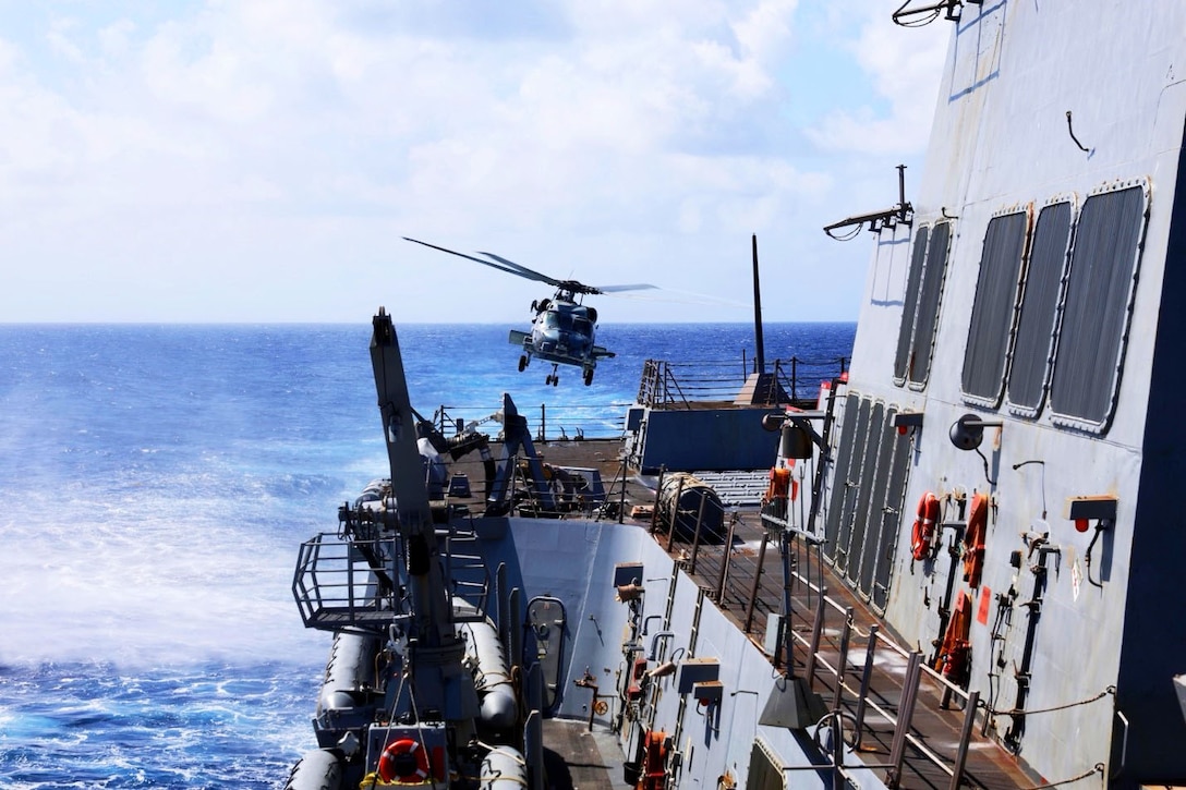 A helicopter lifts off from the deck of a military ship at sea on a cloudy day.