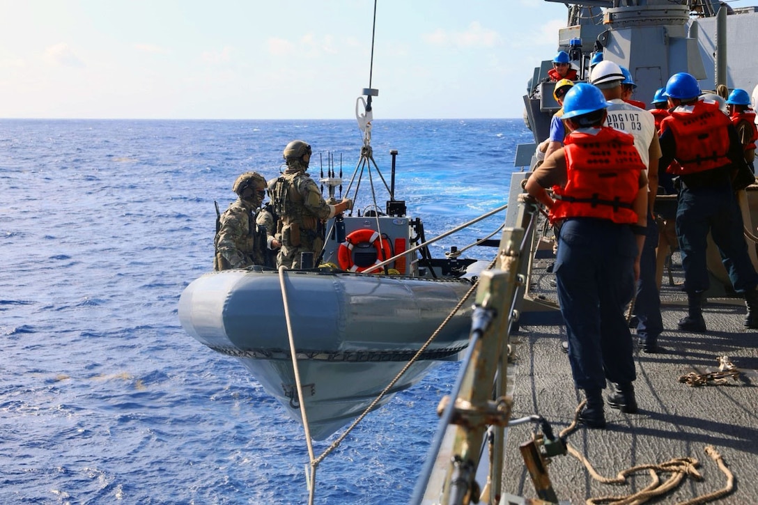 Coast Guardsmen are seen in an inflatable boat attached with ropes to the side of a larger vessel at sea as others on deck watch.
