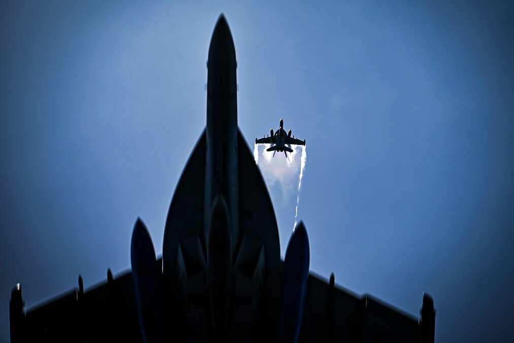 Two jets are seen flying from a view below, with one above the other and contrails visible from its wings.