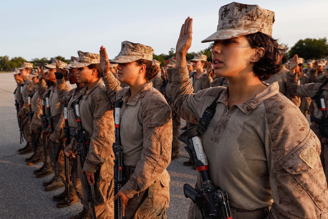 Marine recruits with their right hands raised and carrying rifles around their torsos stand in rows outdoors.