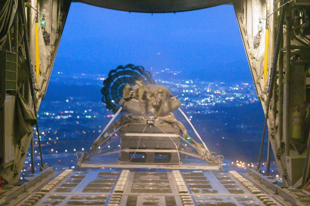 Heavy equipment attached to a parachute sits on the open ramp of an aircraft flying over city lights under a deep blue sky.