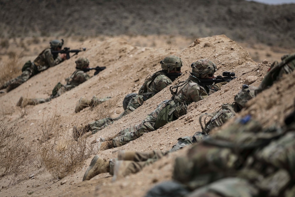 Six soldiers in tactical gear lay on a dirt hill while aiming a weapon.