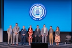 A group in suits and one man in a military uniform stand on a stage with a screen behind.