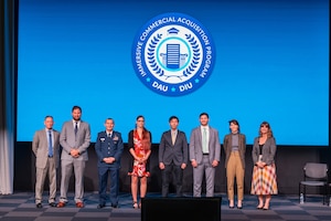 A group in suits and one man in a military uniform stand on a stage with a screen behind.