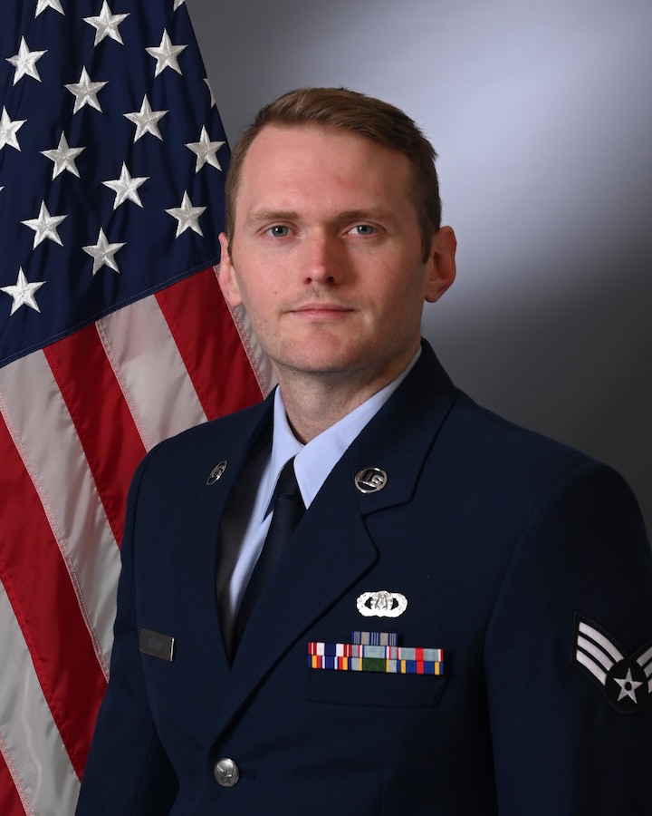 An official photo for the unit website of Senior Airman Michael Gatch standing in front of the American Flag wearing his service dress uniform.