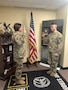 Two Soldiers standing in an office with a flag in the background.