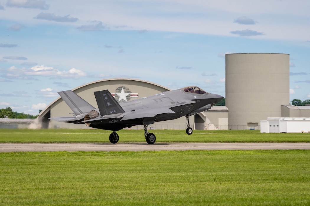 An F-35 crosses in front of beige hangars with the U.S. Air Force logo painted on them