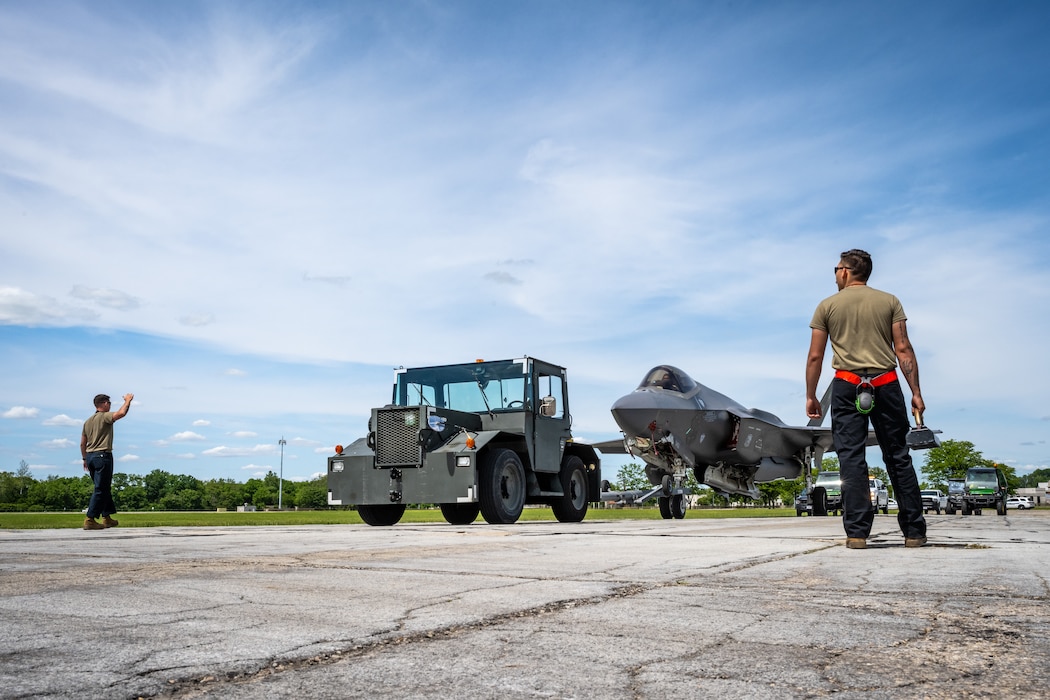 An F-35 behind an Army green 4-wheeled vehicle, two service members in brown tshirts and black pants stand with their back to viewer