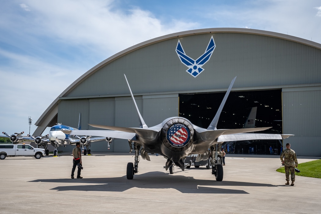 A rear view of a F-35 aircraft in front of a large beige hangar with U.S. Air Force logo painted on front