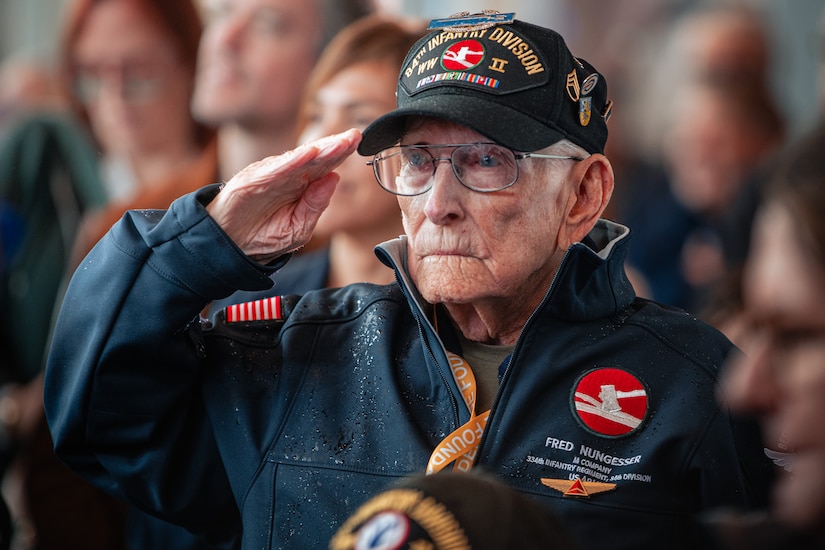 A veteran wearing a hat and jacket salutes while in front of other people.