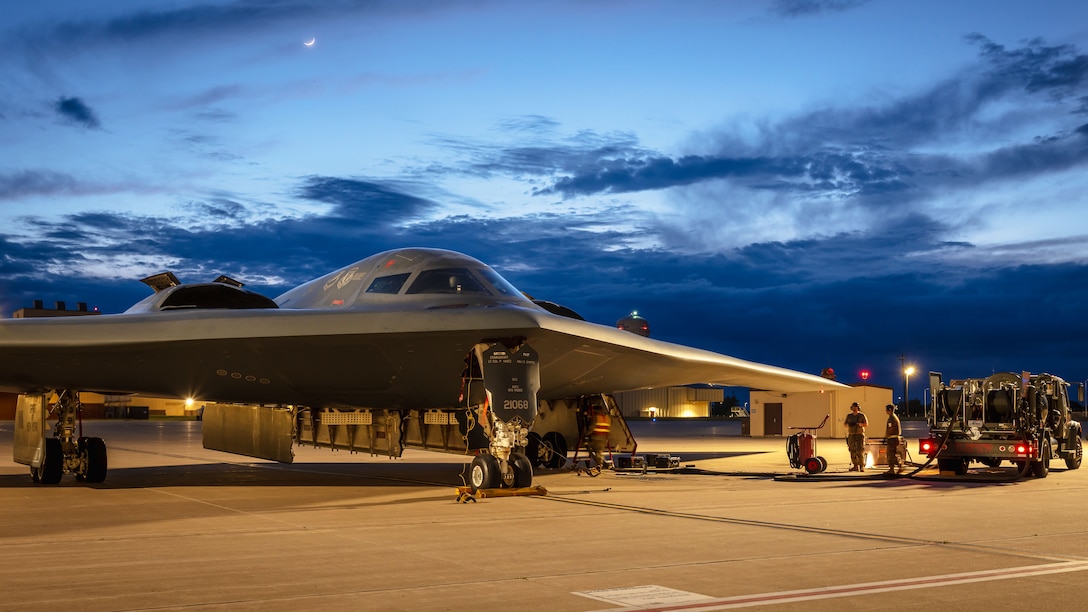Two airmen standing next to a truck on a runway refuel a B-2 aircraft with a crescent moon in a cloudy blue sky in the background.