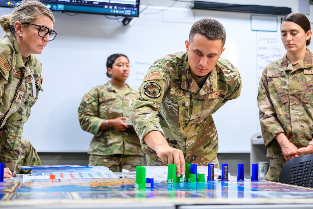 Four people in camouflage uniforms stand around a board on a table with blue and green pieces on it in an indoor, classroom setting.