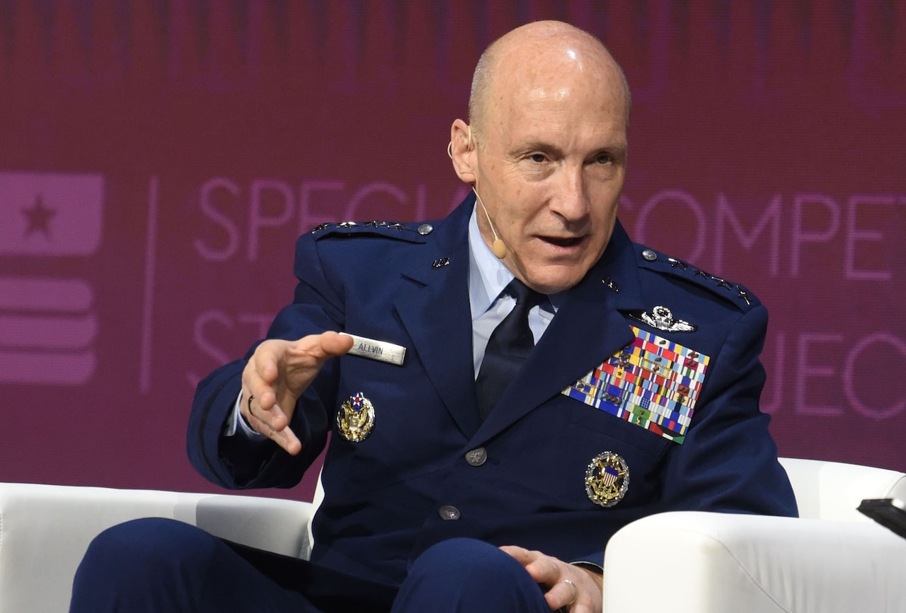 A man wearing a formal military uniform sits in a chair and speaks with a wireless microphone on his head.