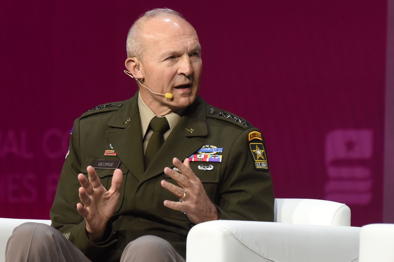 A man wearing a formal military uniform sits in a chair and speaks with a wireless microphone on his head.