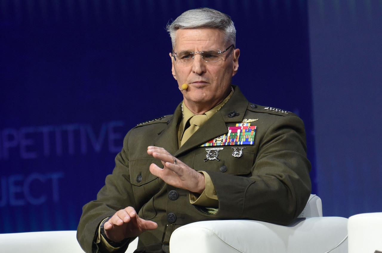 A man wearing a formal military uniform sits in a chair and speaks with a wireless microphone on his head.