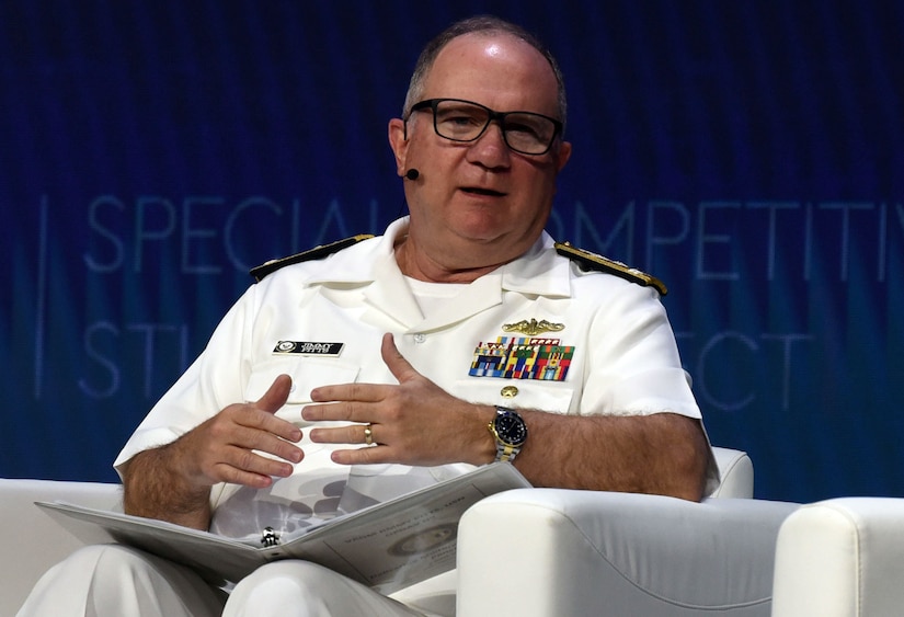 A man wearing a formal military uniform sits in a chair and speaks with a wireless microphone on his head.