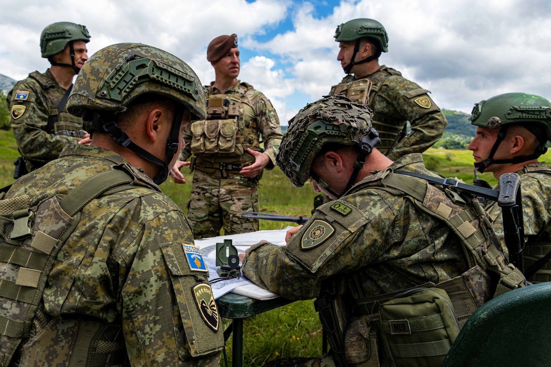 Military personnel in combat uniforms gather around a small table with a compass and papers on it on a grassy field.