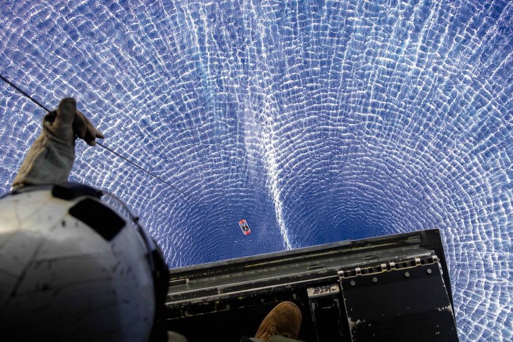 A Marine standing on a helicopter platform and holding a line looks down at a person in floatable device attached to the line as the sea below them ripples.