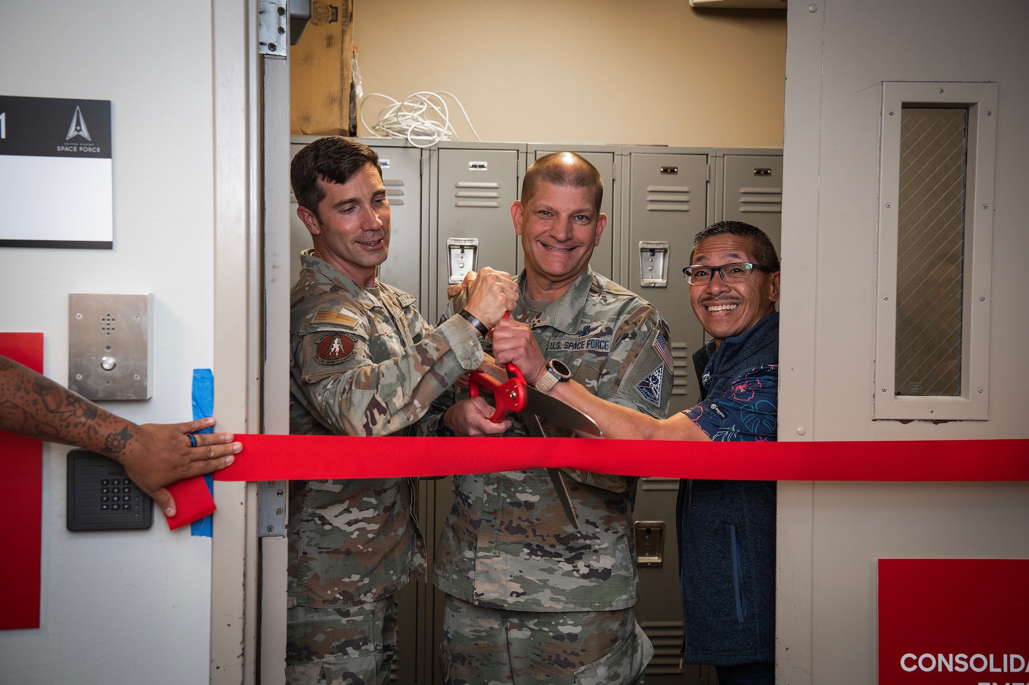 two service members and a civilian hold an oversized pair of scissors for a ribbon cutting ceremony