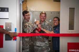 two service members and a civilian hold an oversized pair of scissors for a ribbon cutting ceremony