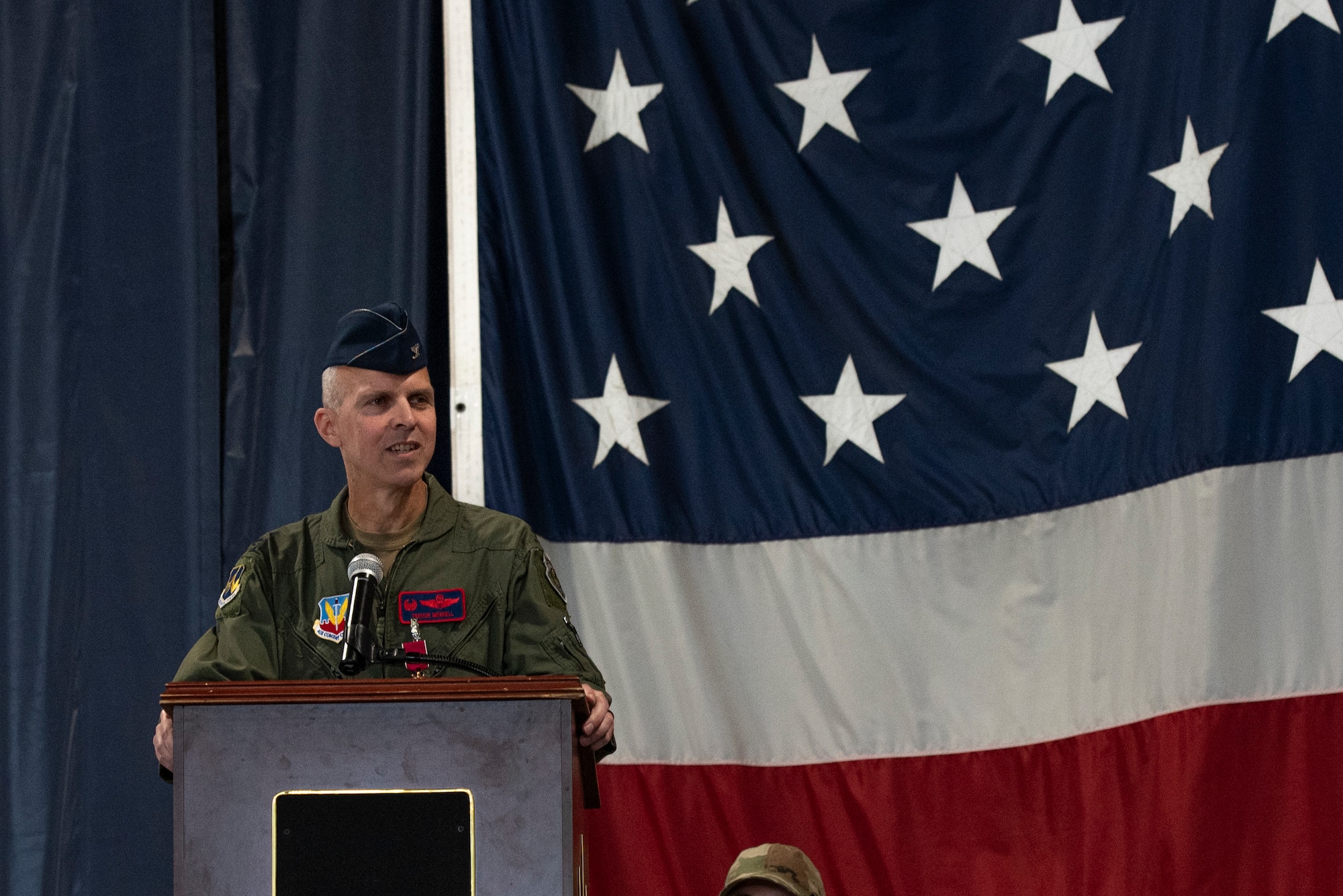 A man in a military flight suit speaks at a podium.