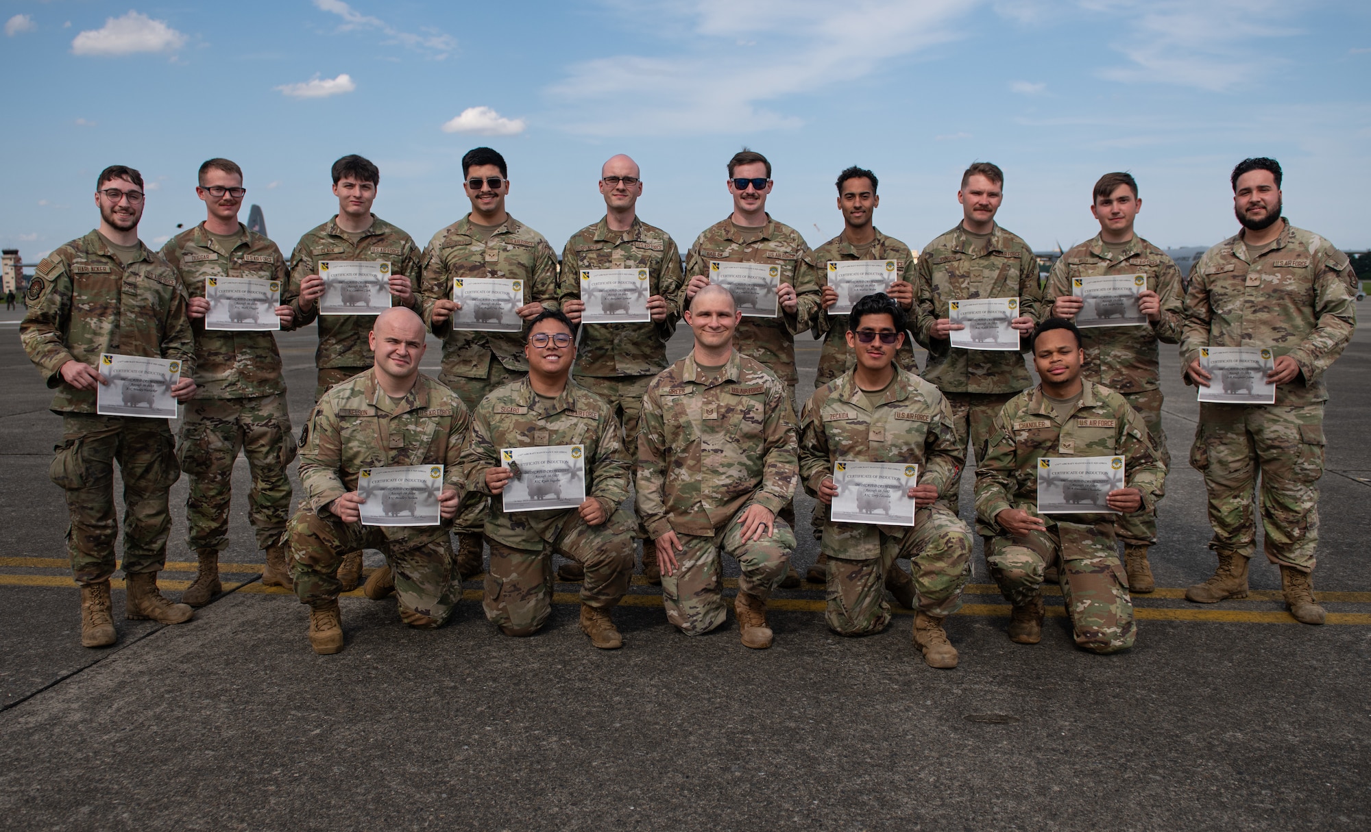 Airmen sit and kneel while holding their certificates in front of them as they pose for a photo on the flightline.