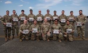 Airmen sit and kneel while holding their certificates in front of them as they pose for a photo on the flightline.
