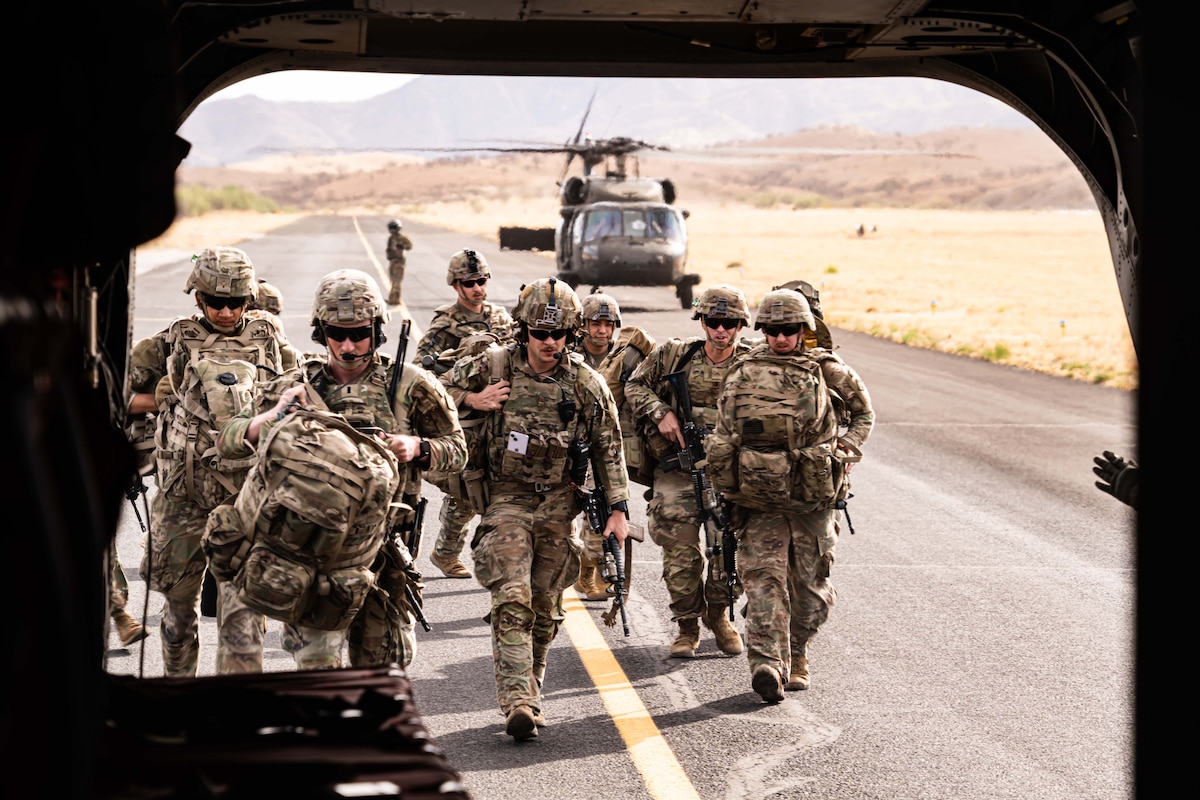 Multiple people wearing camouflage uniforms walk toward the rear of a helicopter while carrying duffle bags and rifles.