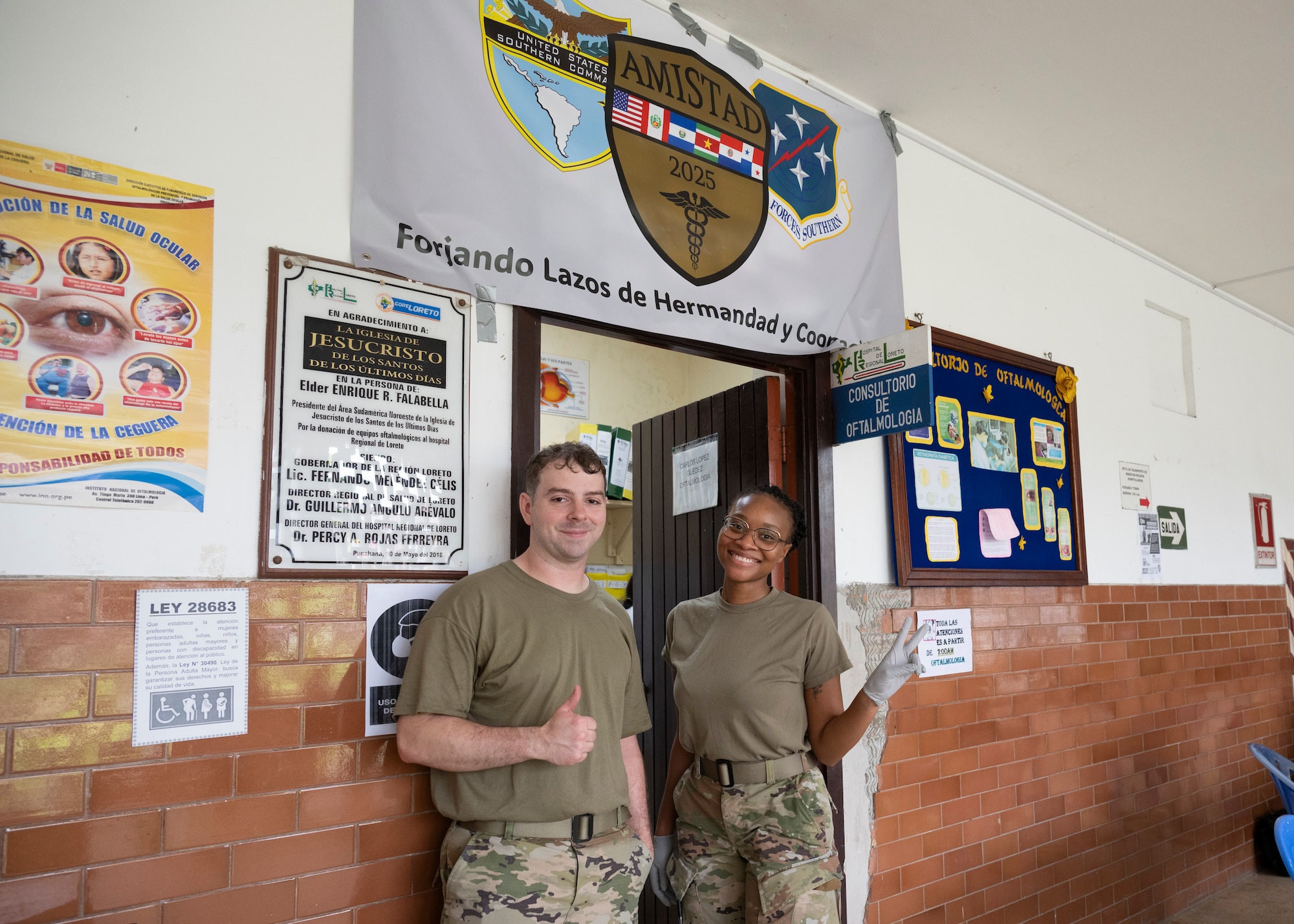 U.S. Air Force Senior Airman Luke Johnson (left) and Senior Airman Erin Bell (right), AMISTAD Optometry Technicians pose outside the optometry clinic door and under the AMISTAD 2025 banner, in Iquitos, Peru, June 2, 2025. AMISTAD enables U.S. and partner nation medics to train and operate in resource constrained environments, building readiness for real-world contingencies. (U.S. Air Force photo by Senior Airman Victoria Edwards)
