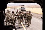 Multiple people wearing camouflage uniforms walk toward the rear of a helicopter while carrying duffle bags and rifles.